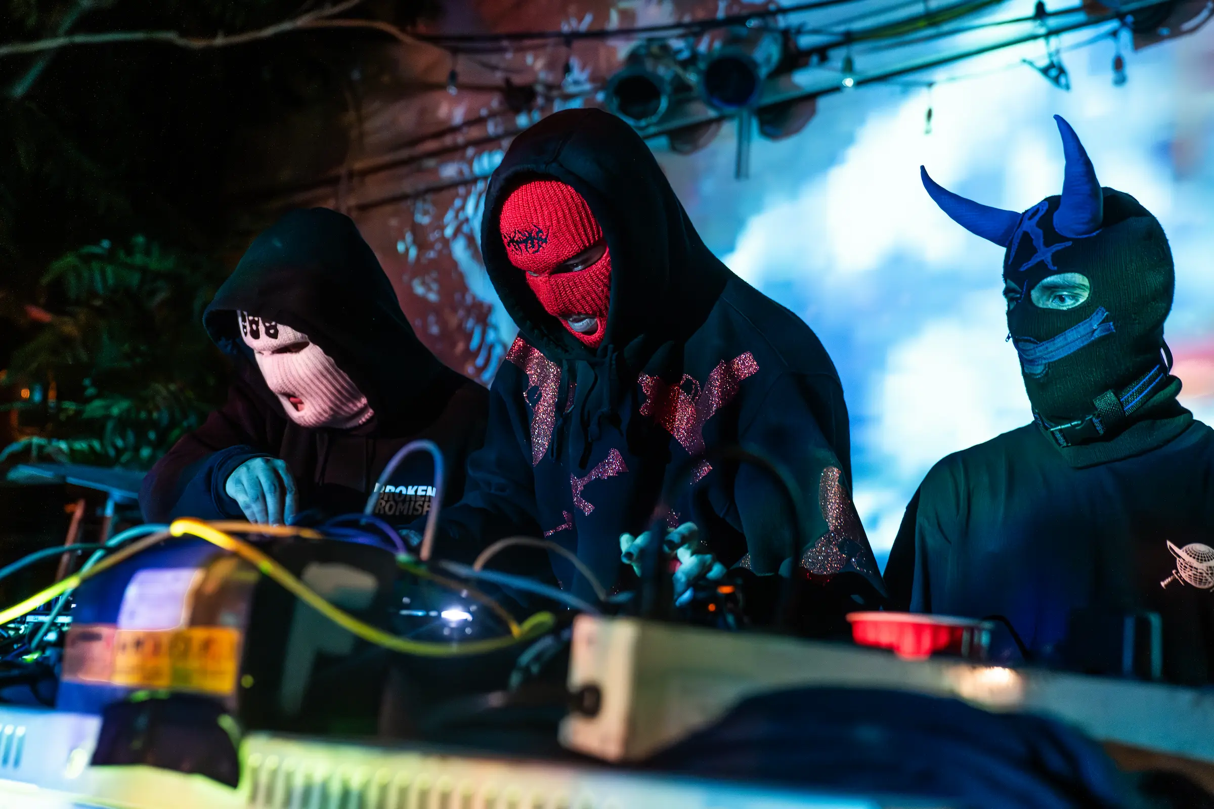 Three masked figures behind a DJ setup under cyan and red stage lighting at night.