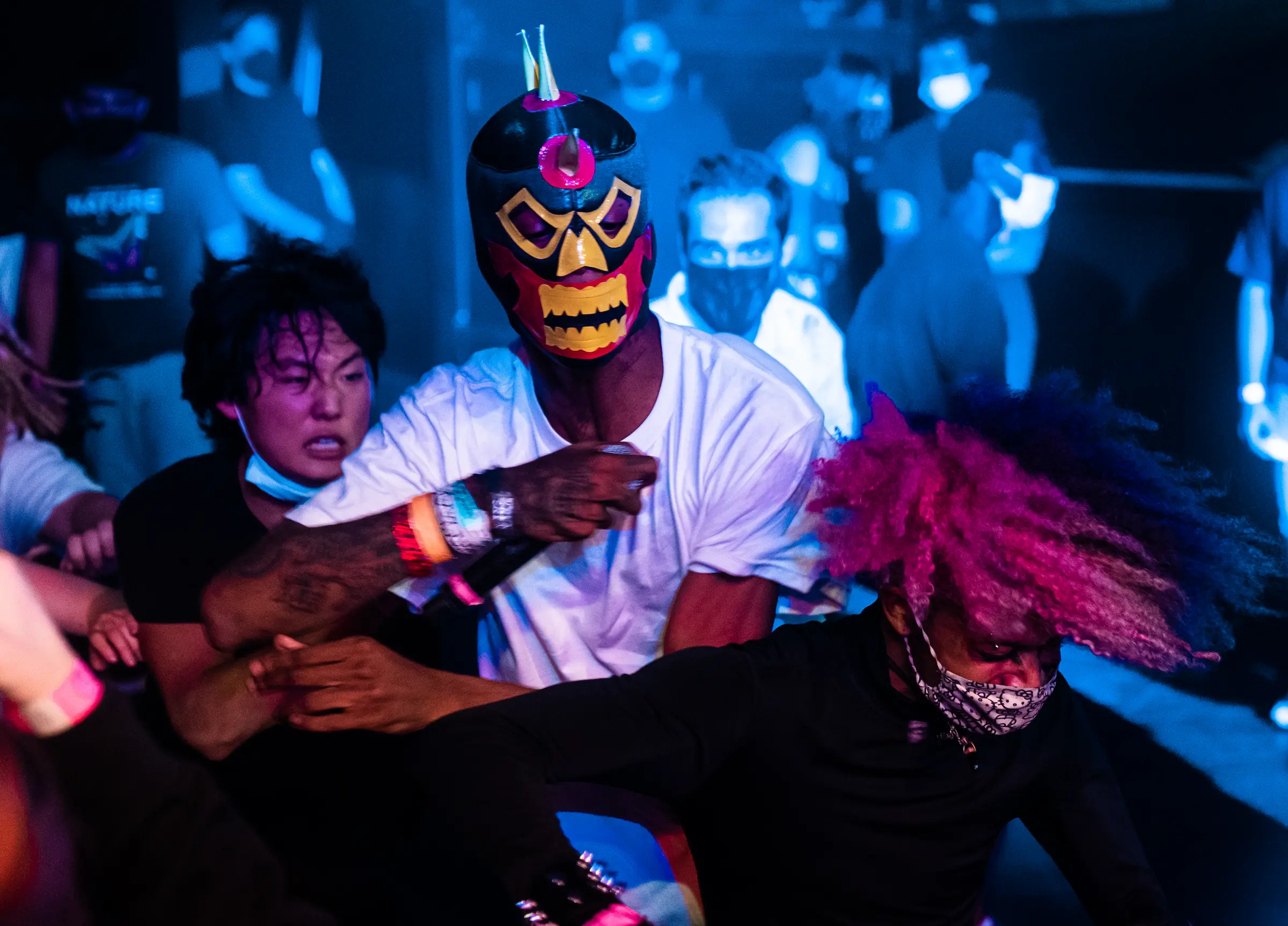 Crowd moment under blue stage light: masked figure and a pink-haired audience member mid-motion.
