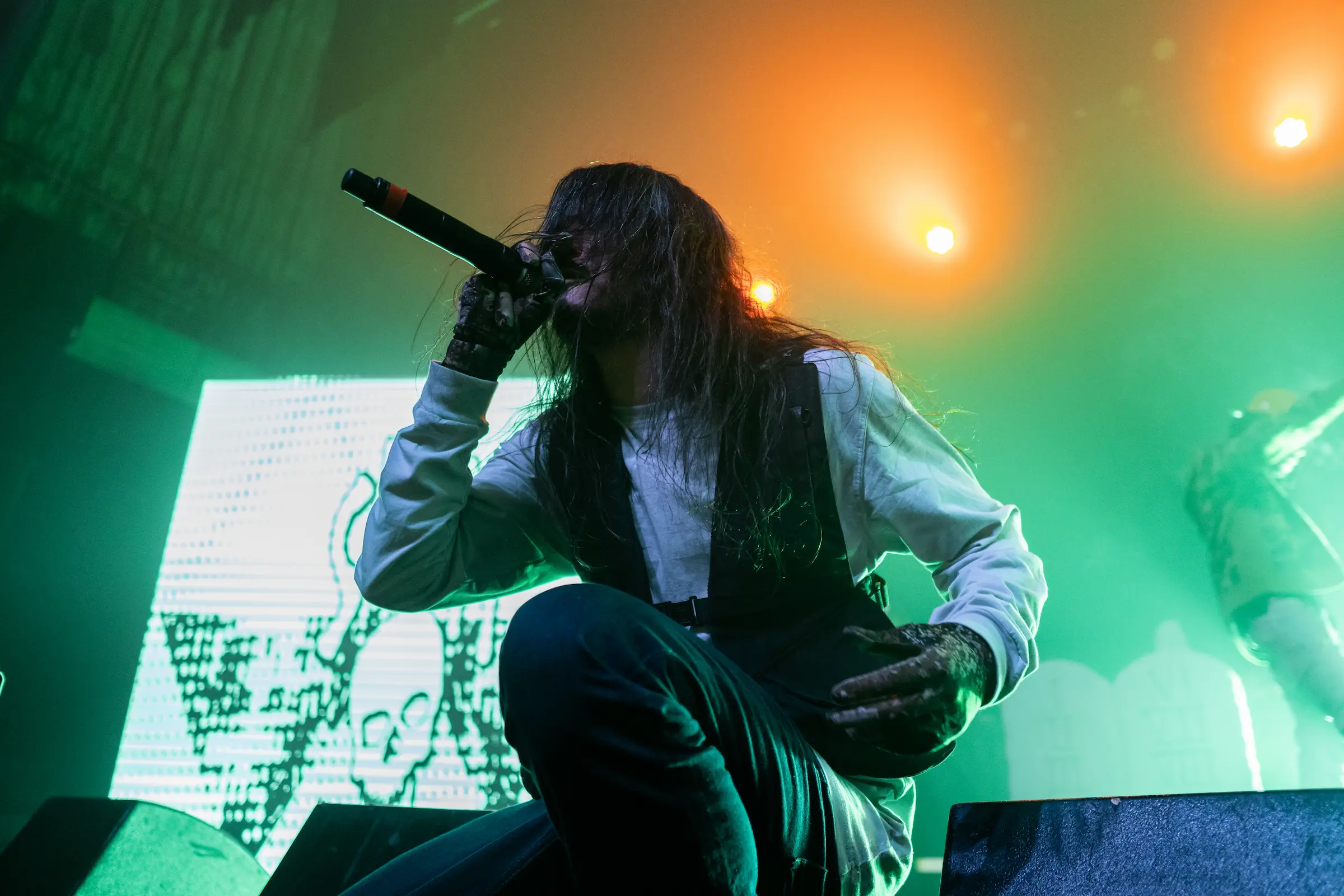 Performer kneeling on stage, microphone close, under green and orange lighting.